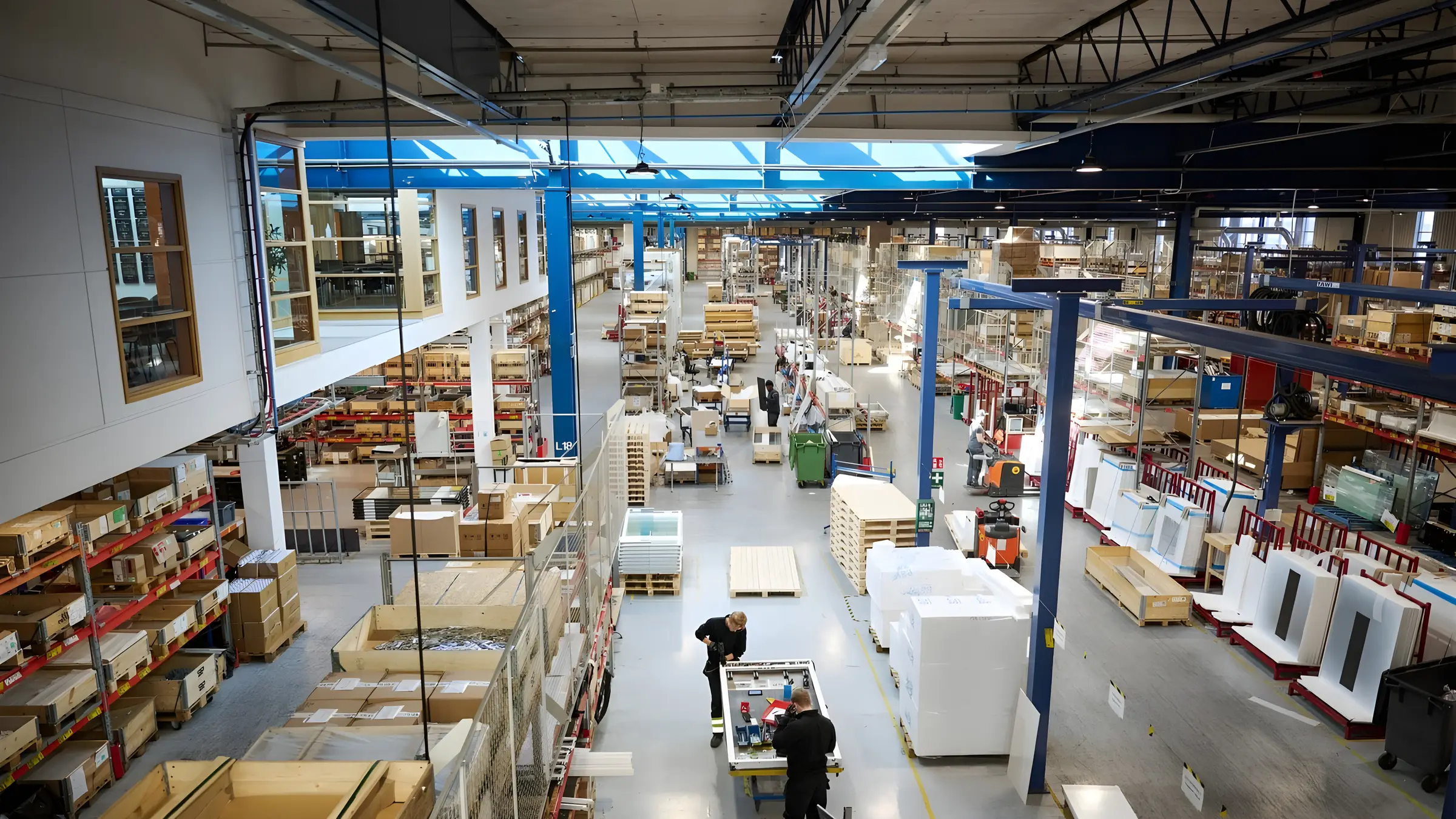 A lift factory's assembly hall seen from above, with parts stacked on racking along both walls and two workers at a bench in the centre.